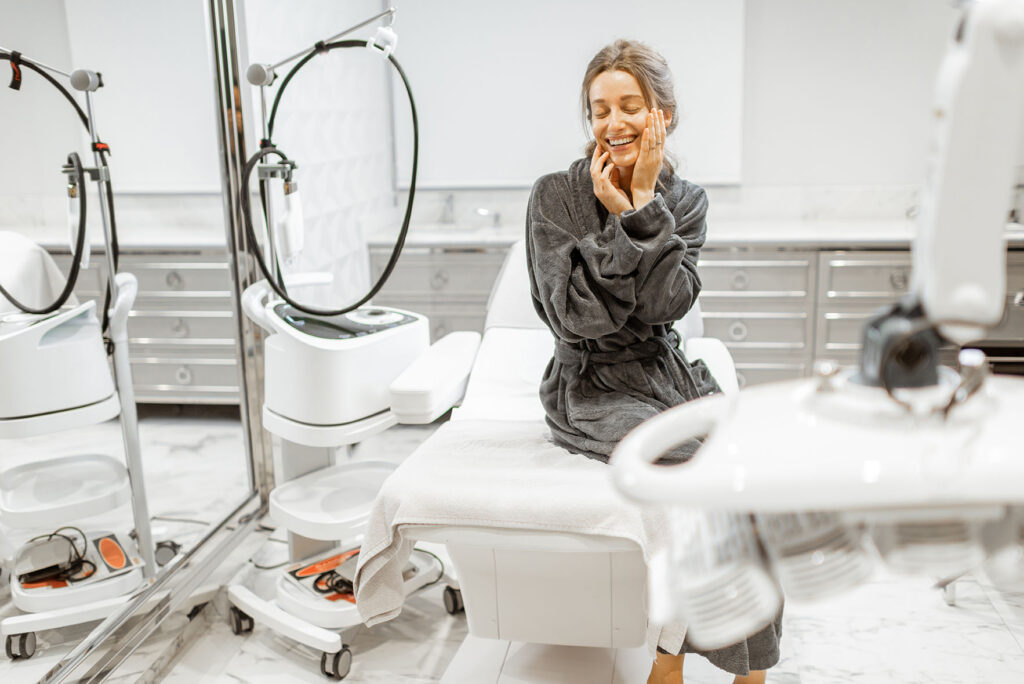 woman in a dark robe sitting on a medical chair in a modern treatment room surrounded by skincare equipment