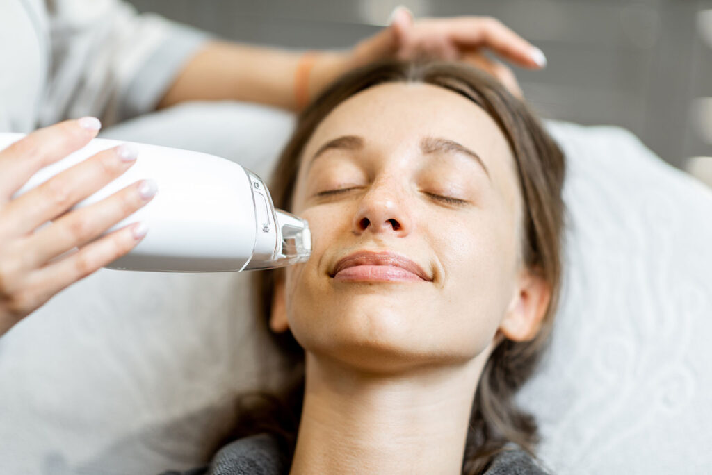 woman lying on a treatment bed with eyes closed while a clinician uses a white handheld laser device on her cheek