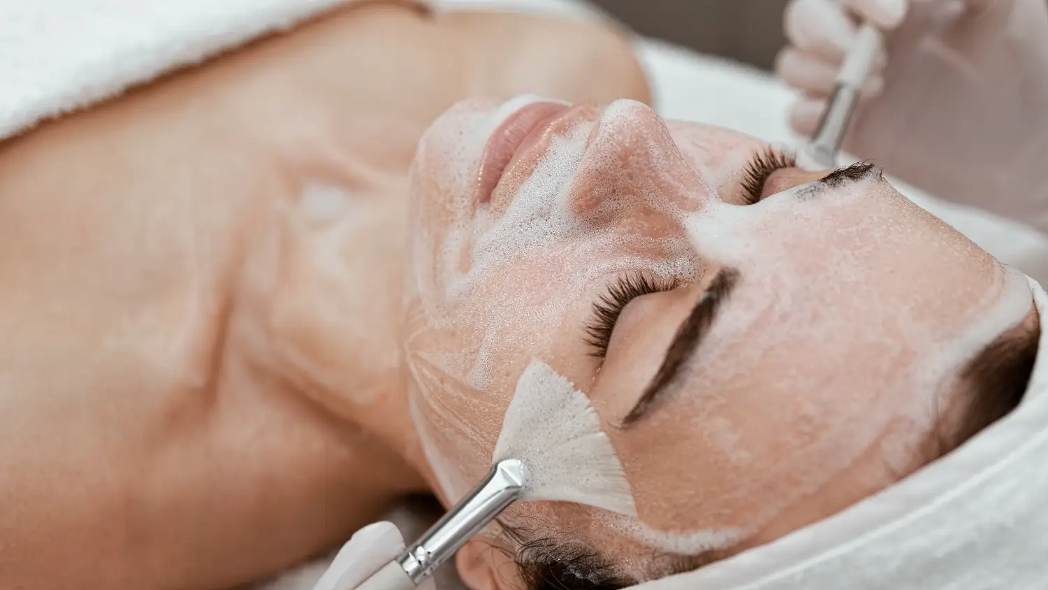 Woman lying down with foaming cleanser applied while brushes touch her cheeks