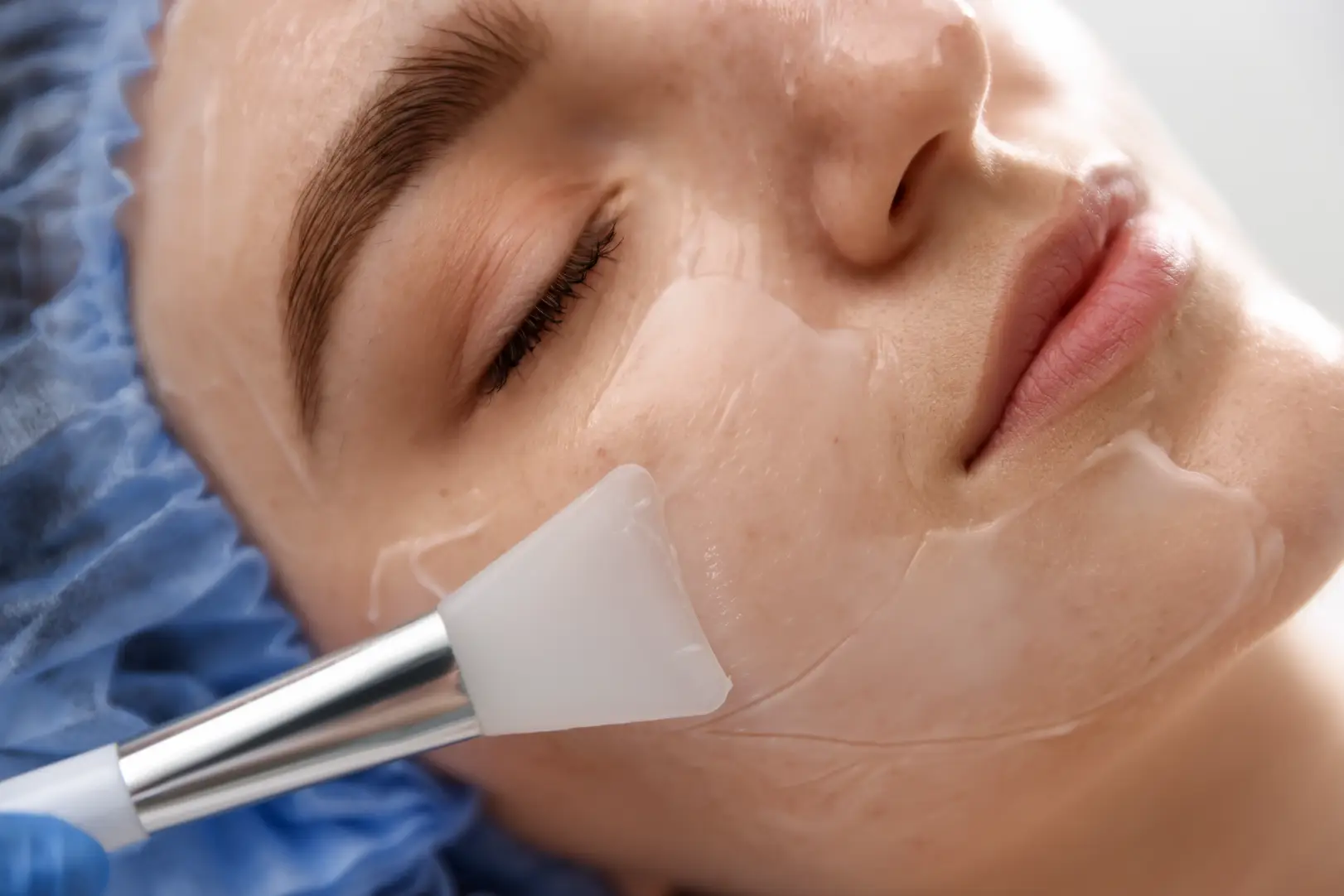 Close-up of a woman with eyes closed as a brush applies a clear facial solution to her cheek.